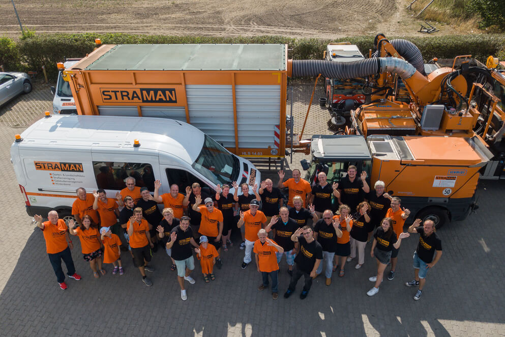Gruppe von Mitarbeitenden steht vor orangefarbenen Nutzfahrzeugen und winkt in die Kamera.