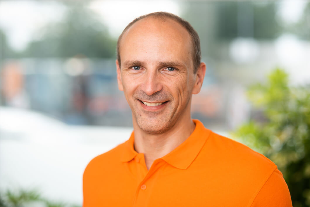 Dirk Borrmann – STRAMAN GmbH Porträt eines freundlich lächelnden Mannes mit kurzen braunen Haaren und orangefarbenem Poloshirt vor hellem Hintergrund mit Grünpflanzen.