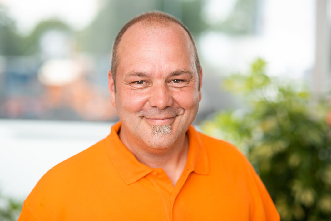 Porträt eines freundlich lächelnden Mannes mit sehr kurzen braunen Haaren, grauem Kinnbart und orangefarbenem Poloshirt vor hellem Hintergrund.