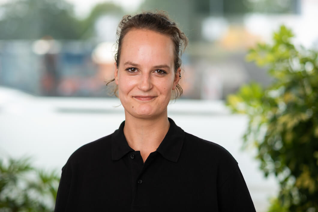 Porträt einer freundlich lächelnden Mitarbeiterin mit hochgebundenen braunen Haaren und schwarzem Poloshirt vor einem hellen Hintergrund.