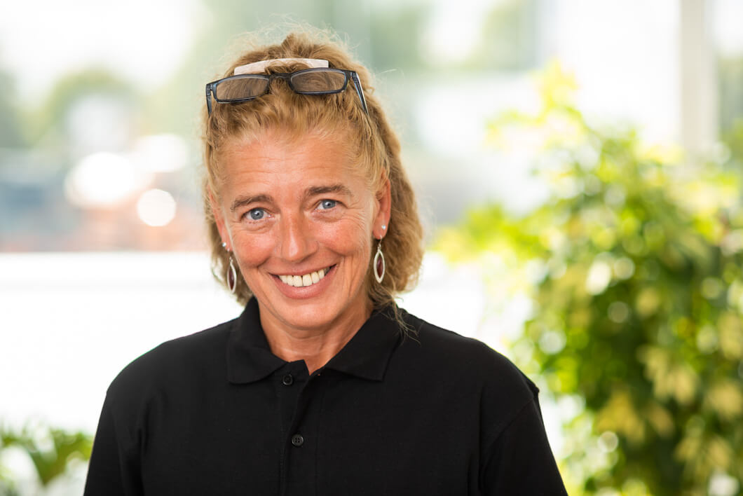 Porträt einer freundlich lächelnden Frau mit blonden Haaren, hochgesteckter Brille und schwarzem Poloshirt vor hellem Hintergrund mit Grünpflanzen.