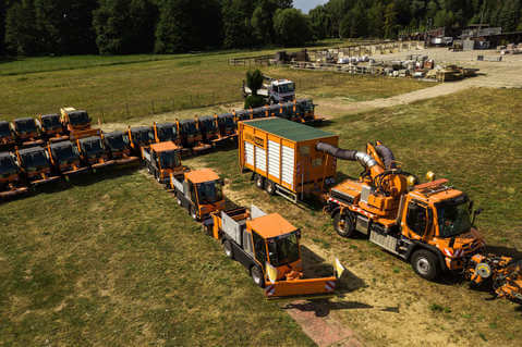 Mehrere orange Arbeitsfahrzeuge stehen in einer Reihe neben einem großen Saugfahrzeug mit Anhänger.