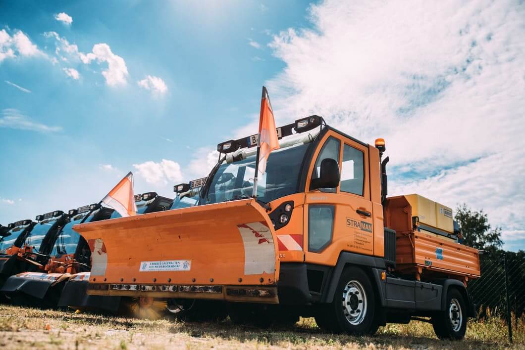 STRAMAN Winterdienstfahrzeuge mit Schneepflug Orangefarbenes STRAMAN Winterdienstfahrzeug mit Schneepflug und Streuaufbau vor einer Flotte weiterer Räumfahrzeuge.