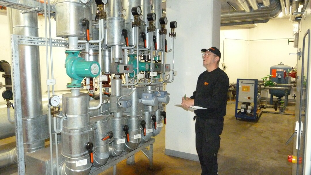 Mitarbeiter bei der Kontrolle einer technischen Heizungs- und Pumpenanlage mit Rohrleitungen und Messgeräten im Gebäudemanagement.