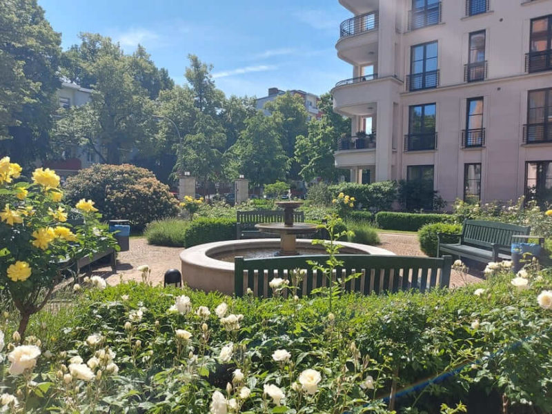 Grünanlagenpflege Berlin _ STRAMAN Gepflegte Gartenanlage mit Brunnen, blühenden Rosen, Hecken und Sitzbänken vor einem Wohngebäude