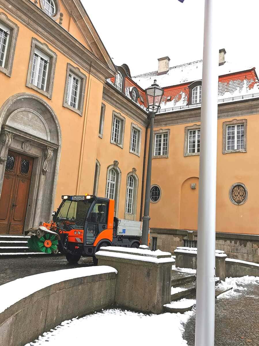 STRAMAN Winterdienst Orangefarbenes STRAMAN-Winterdienstfahrzeug mit Kehrbürste bei der Schneeräumung vor einem historischen Gebäude.