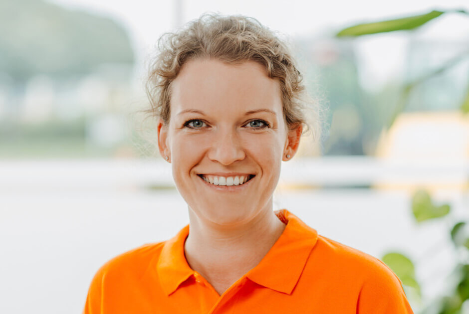 Alexandra Kuhn -STRAMAN GmbH Porträt einer lächelnden STRAMAN-Mitarbeiterin im orangefarbenen Poloshirt vor einem hellen, unscharfen Hintergrund.