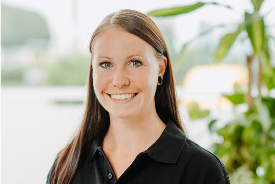 Franziska Henkel – STRAMAN GmbH Berlin Porträt einer lächelnden STRAMAN-Mitarbeiterin im schwarzen Poloshirt vor einem hellen Hintergrund mit grünen Pflanzen.