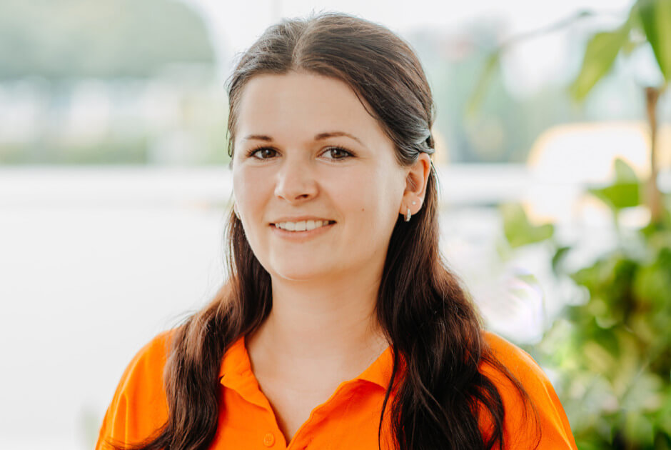 Porträt einer lächelnden STRAMAN-Mitarbeiterin im orangefarbenen Poloshirt vor einem hellen, begrünten Hintergrund.