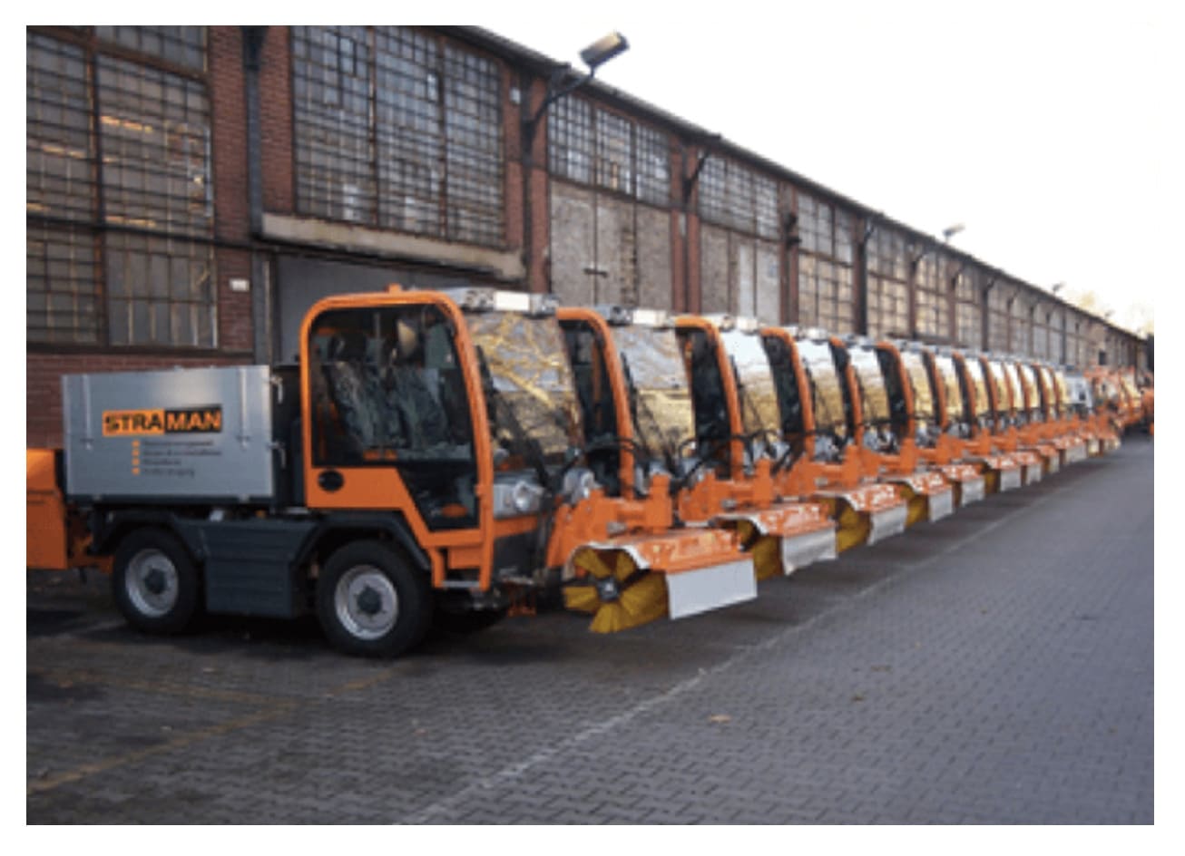 Mehrere orangefarbene STRAMAN Kehrmaschinen stehen vor einer Industriehalle in einer Reihe.
